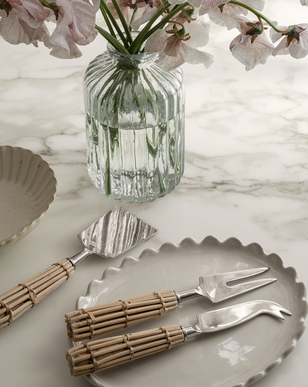 A marble table set with scalloped-edge beige plates, a Verlice Glass Cylinder Vase filled with pink flowers, and three cheese utensils with silver metal heads and woven rattan handles arranged neatly on a plate.