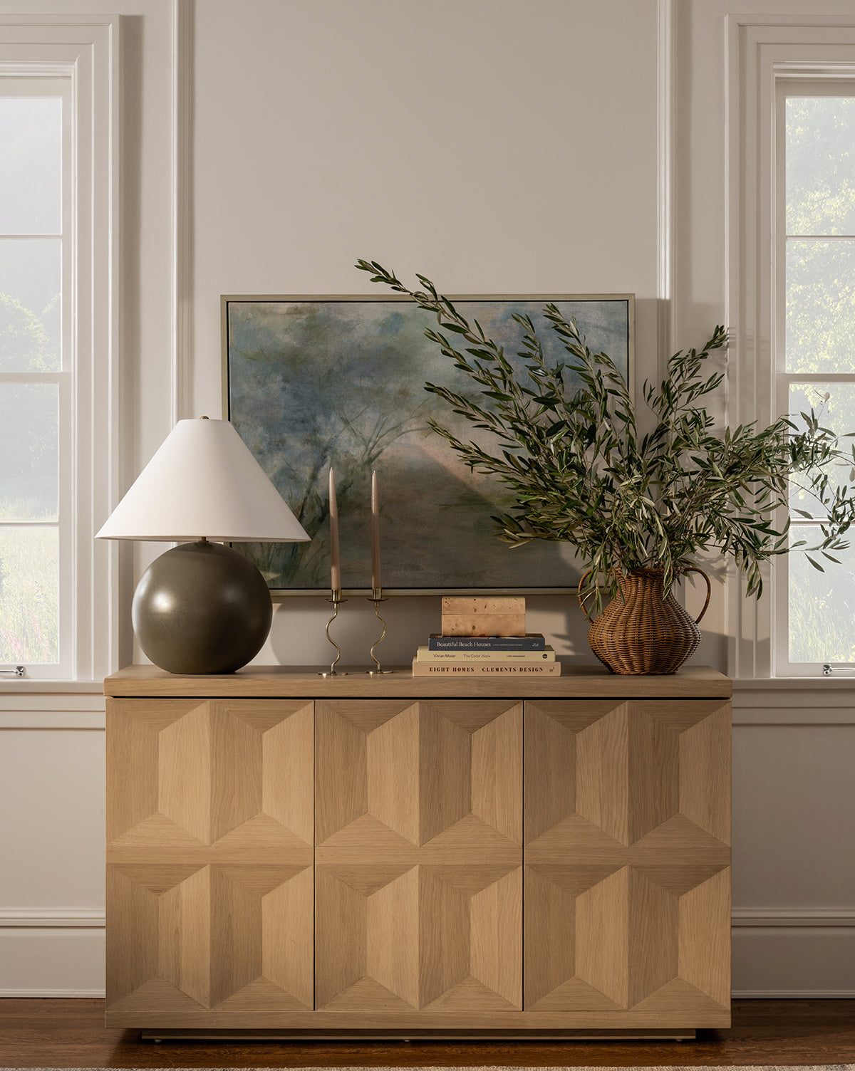 Between two windows sits a wooden sideboard with a geometric pattern, topped with a table lamp, two candlesticks, stacked books, a framed giclee print titled Fading, and a vase of leafy branches.