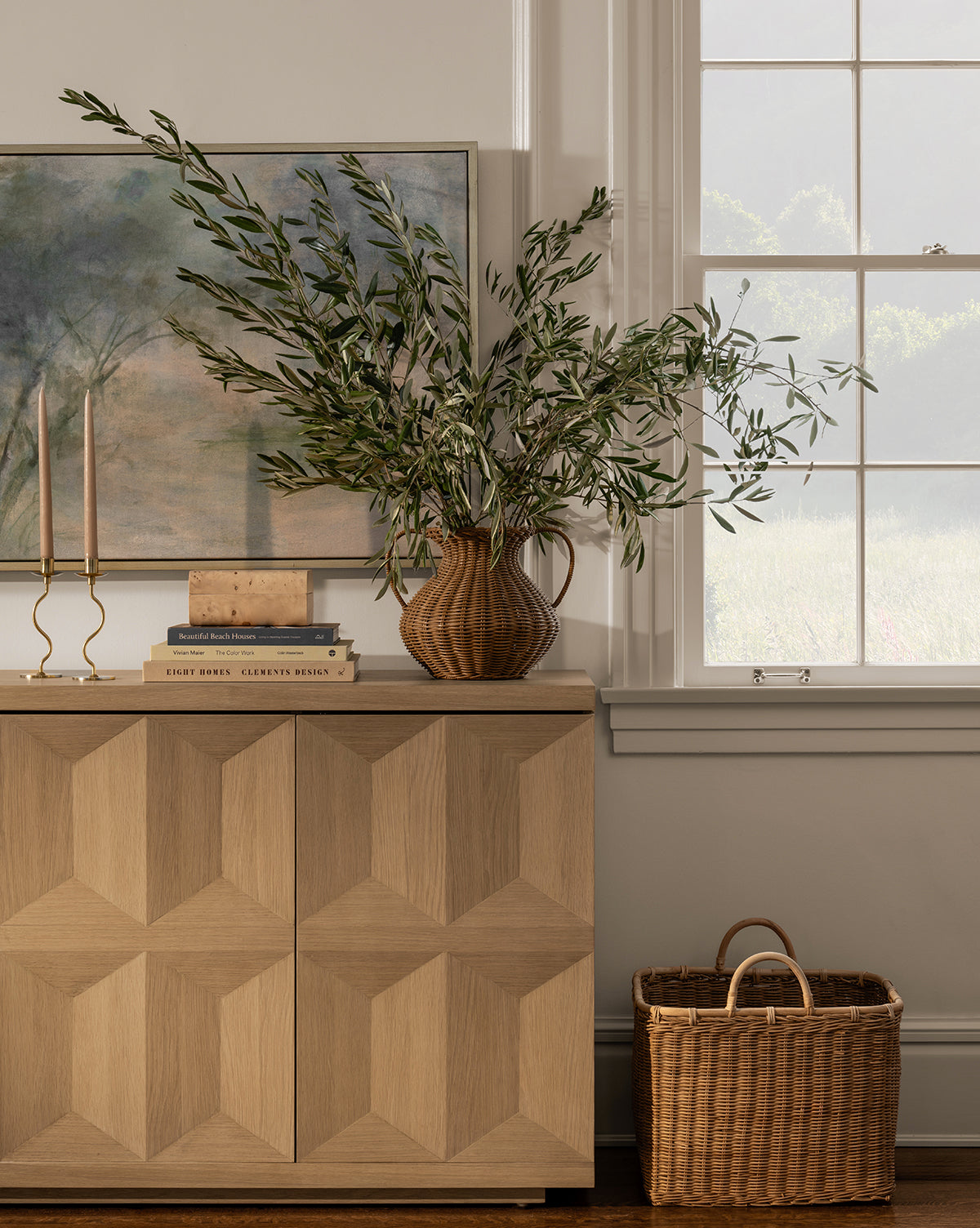 A wooden cabinet with geometric patterns displays the Antonia Woven Vase filled with leafy branches, stacked books, and two gold candlesticks. A basket rests on the floor, with a window and framed painting in the background.