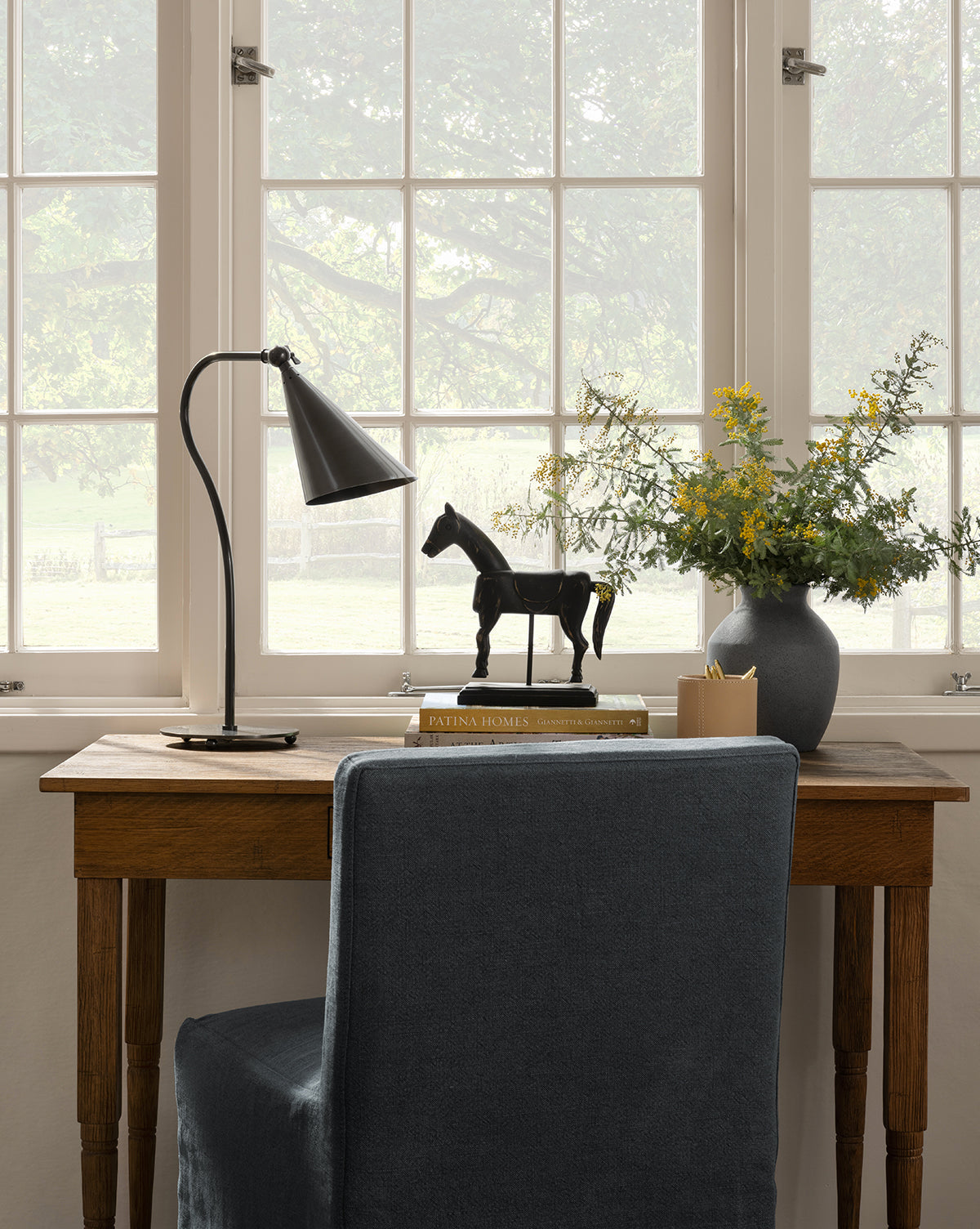 A wooden desk with a gray chair faces large windows. On the desk sit a Lupe Table Lamp, books, a black horse figurine, a vase of yellow flowers, and a pencil-filled cup. Trees can be seen outside through the windows by McGee & Co.