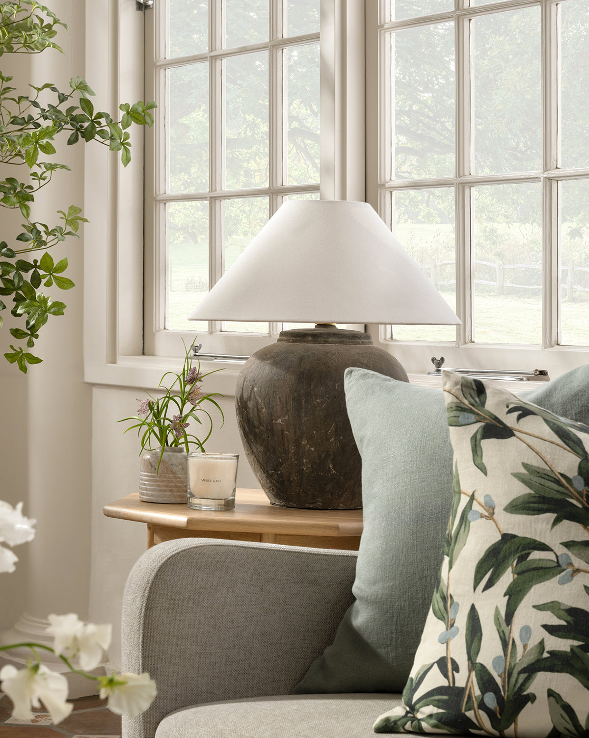 A cozy corner with a sofa, leafy pillow, light blue cushion, and wooden side table topped with a rustic lamp, Stinson Marble Bud Vase, small plant, and candle—set by large sunlit windows framing green outdoor foliage, by McGee & Co.