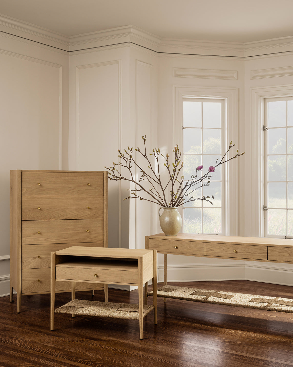 A bright room with wood floors features the Dana White Oak Nightstand, a minimalist light wood piece, along with a tall dresser and console table, set against white paneled walls and large windows, by McGee & Co.