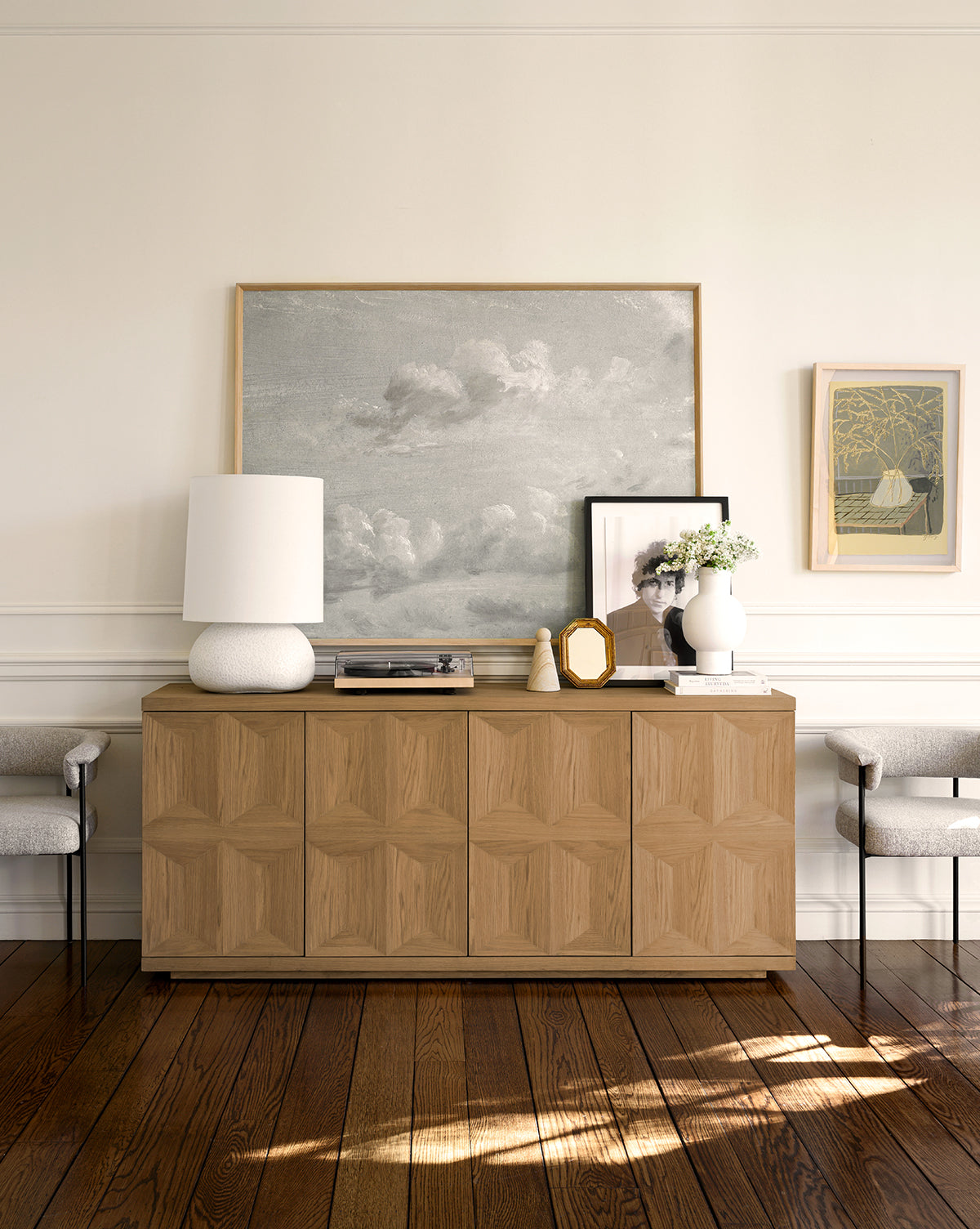 The Beacon Sideboard features a neutral finish and geometric patterns, providing ample storage. Pair it with a white lamp, framed photos, flowers, art above, and two upholstered chairs for a stylish look against a crisp white wall by McGee & Co.