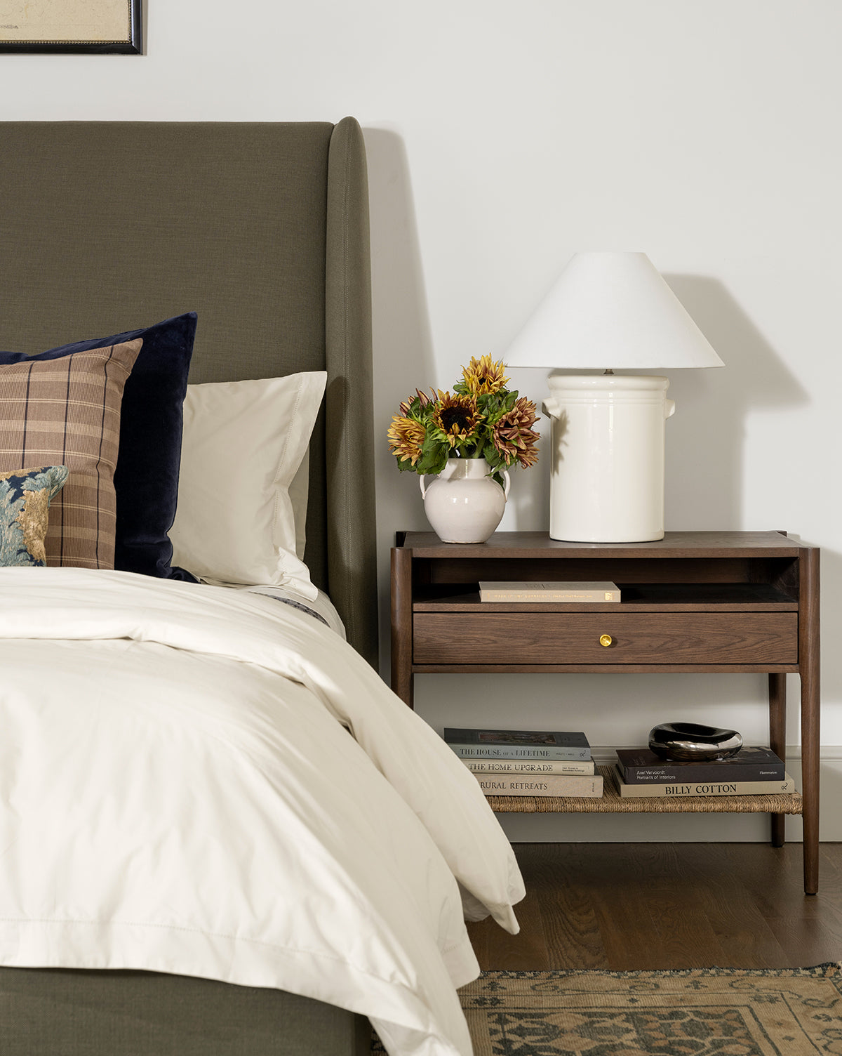 A cozy bedroom scene with a green upholstered bed, white bedding, and a Dana Walnut Oak Nightstand with brass hardware. On the nightstand are a white lamp, a sunflower vase, and stacked books on the lower shelf by McGee & Co.