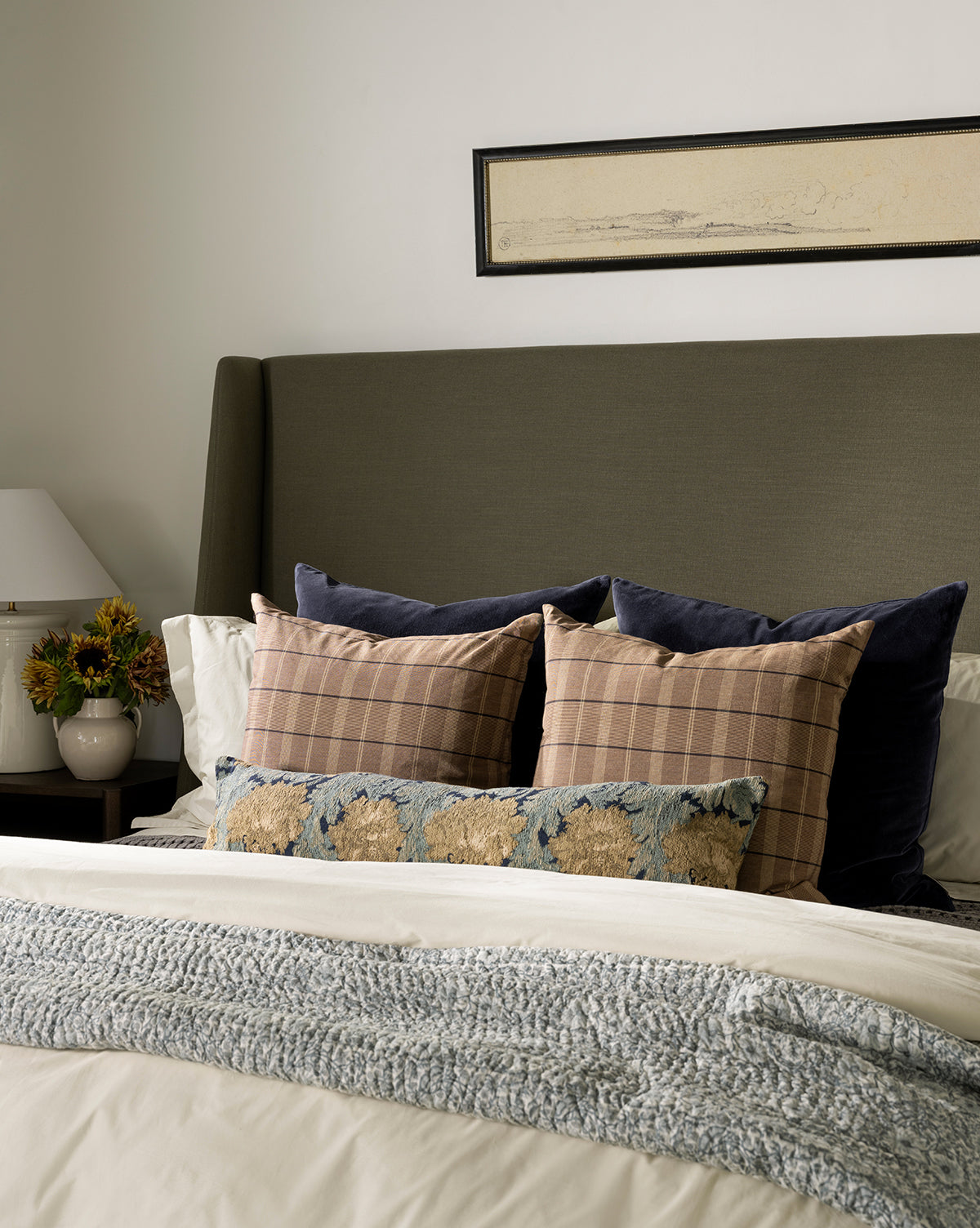 A neatly made bed with layered pillows, including a decorative pillow in the Bellevue Velvet Pillow Cover, a textured throw blanket, a potted plant and lamp on the bedside table, and rectangular framed artwork above the headboard, by McGee & Co.