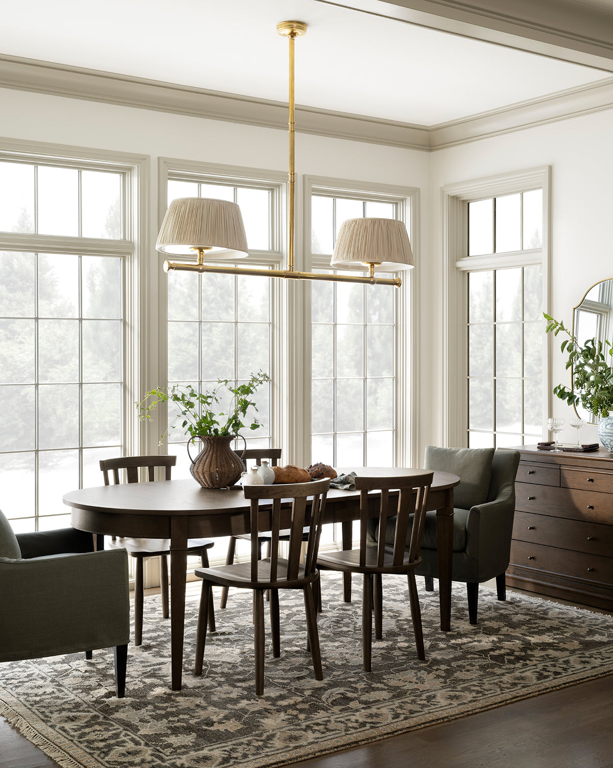 A bright dining room with large windows, a round dark wood table surrounded by Hemingway Chairs, a patterned rug, green armchairs, a vase of greenery, a sideboard, and a modern gold chandelier with two lampshades, by McGee & Co.