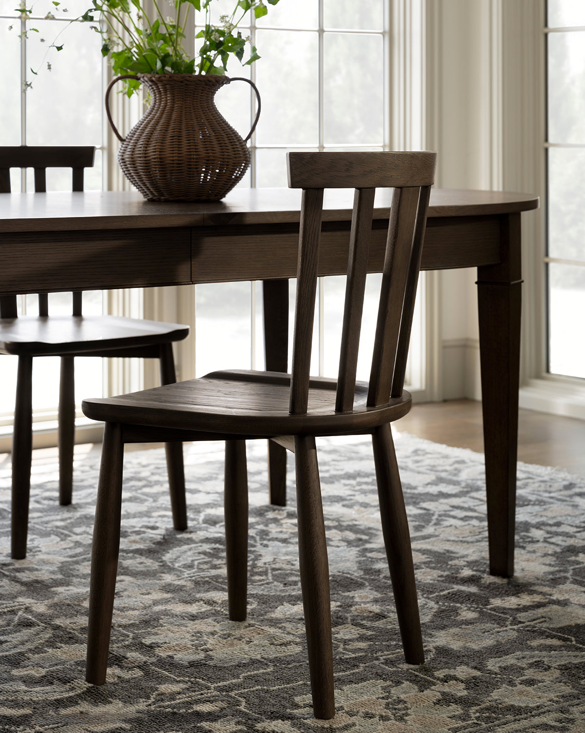 A Hemingway Chair with a slatted back sits on a patterned rug beside a matching table and wicker vase with greenery, illuminated by natural light from large windows, by McGee & Co.