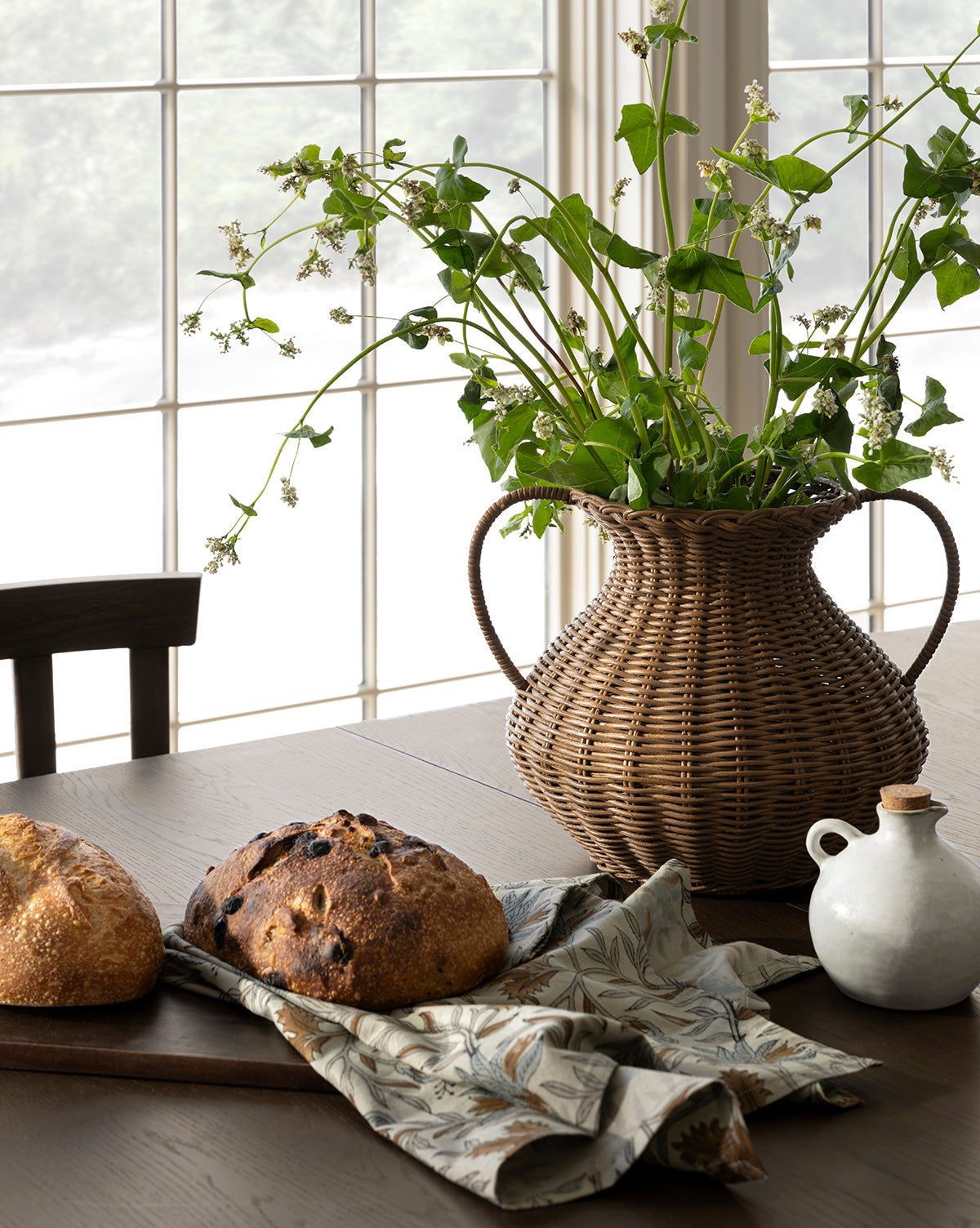 A wooden table is set with two loaves of bread on a cutting board, a patterned cloth, a white ceramic jug, and the Antonia Woven Vase filled with green leafy branches, in front of large sunlit windows, by McGee & Co.