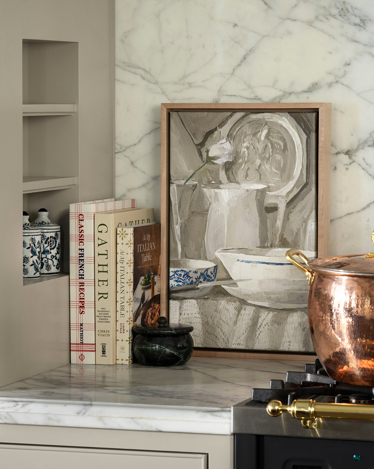 A kitchen countertop displays the "Classic French Recipes" cookbook, a decorative jar, marble mortar and pestle, framed still-life art, and a copper pot on the stove, all set against a marble backsplash, by McGee & Co.