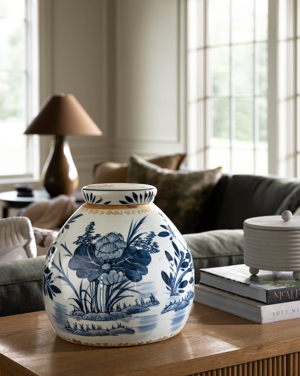 A Chinoiserie Ginger Jar with blue and white hand-painted floral patterns sits on a wooden table in a bright living room, complemented by a green sofa, books, and large windows in the background, by McGee & Co.