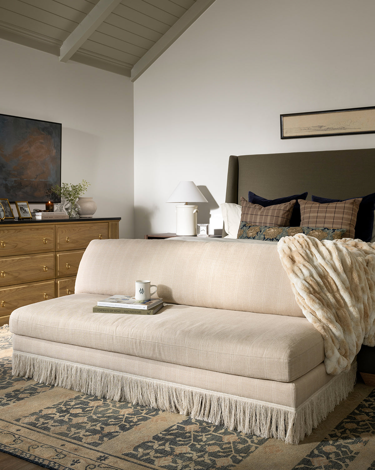 A cozy bedroom showcases a beige upholstered bench with fringe, a soft fur throw, the M&Co Diner Mug, books, a wooden dresser, framed art, and a patterned rug on the floor, by McGee & Co.