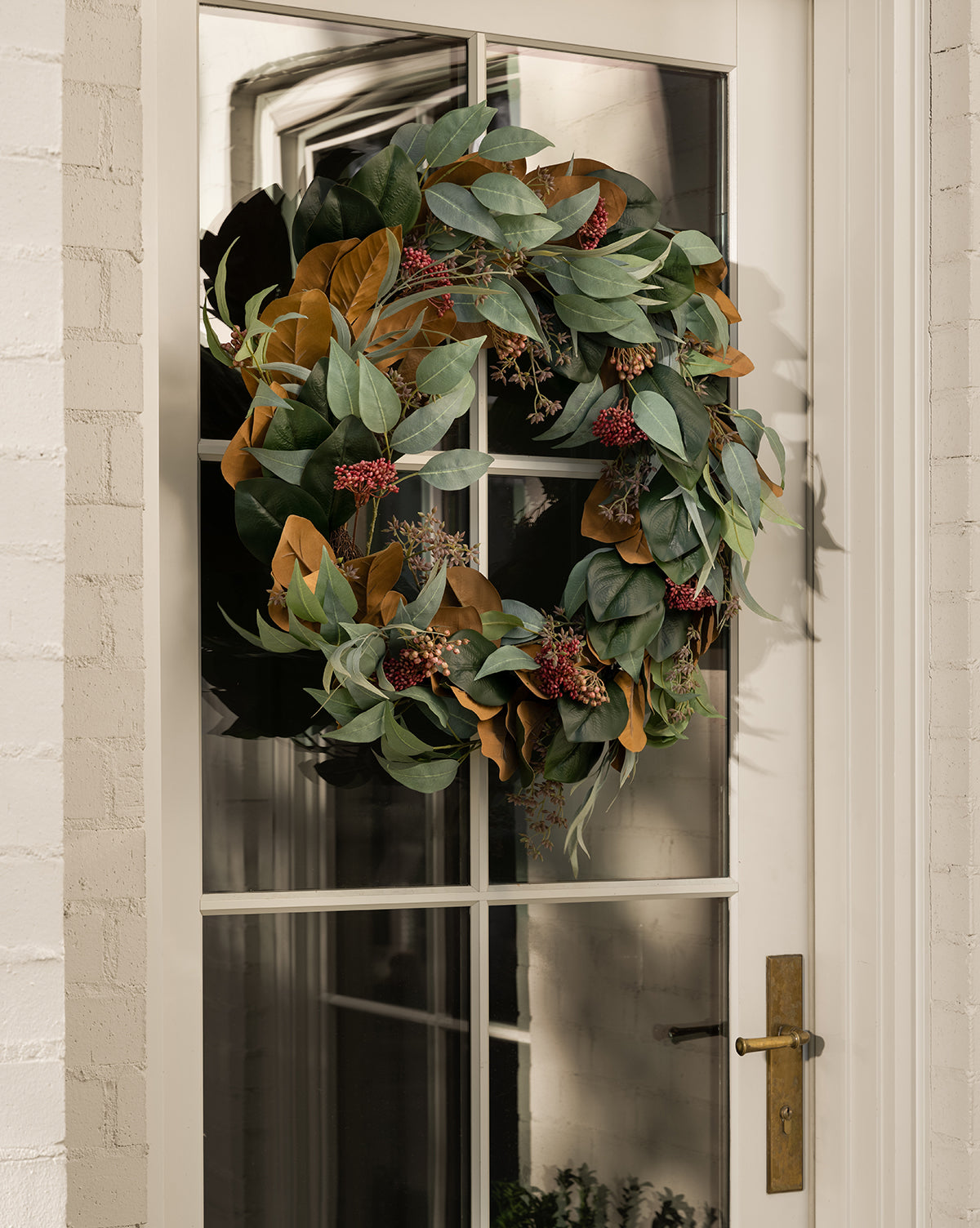 A 30 Faux Magnolia & Eucalyptus Wreath hangs on a glass-paneled door with a brass handle, reflecting light and the white brick exterior.
