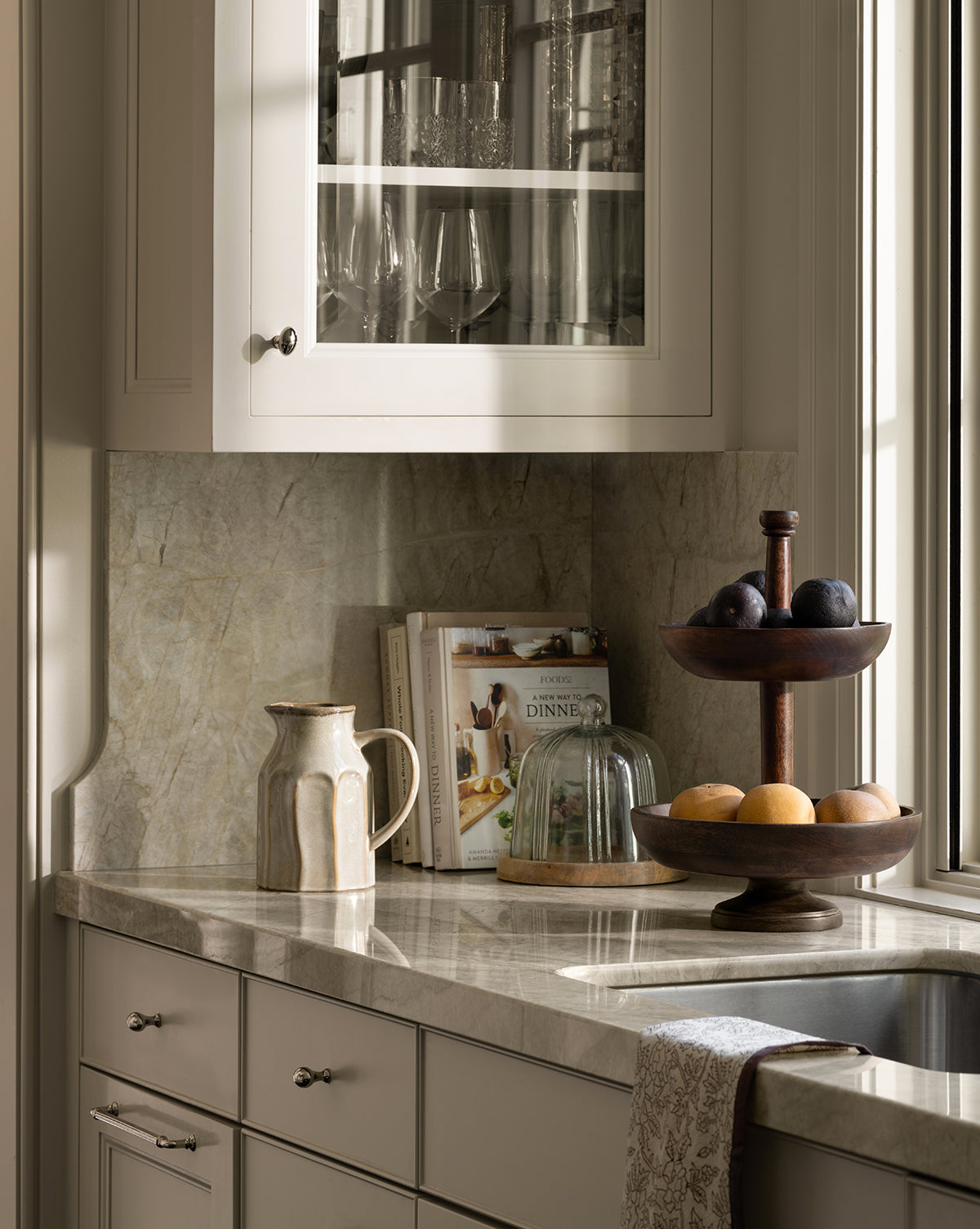 A sunlit kitchen counter features the Winward Tiered Pedestal holding plums and lemons, alongside a sculpted ceramic pitcher, cookbooks, and a glass-covered dish beneath a cabinet displaying glassware by McGee & Co.