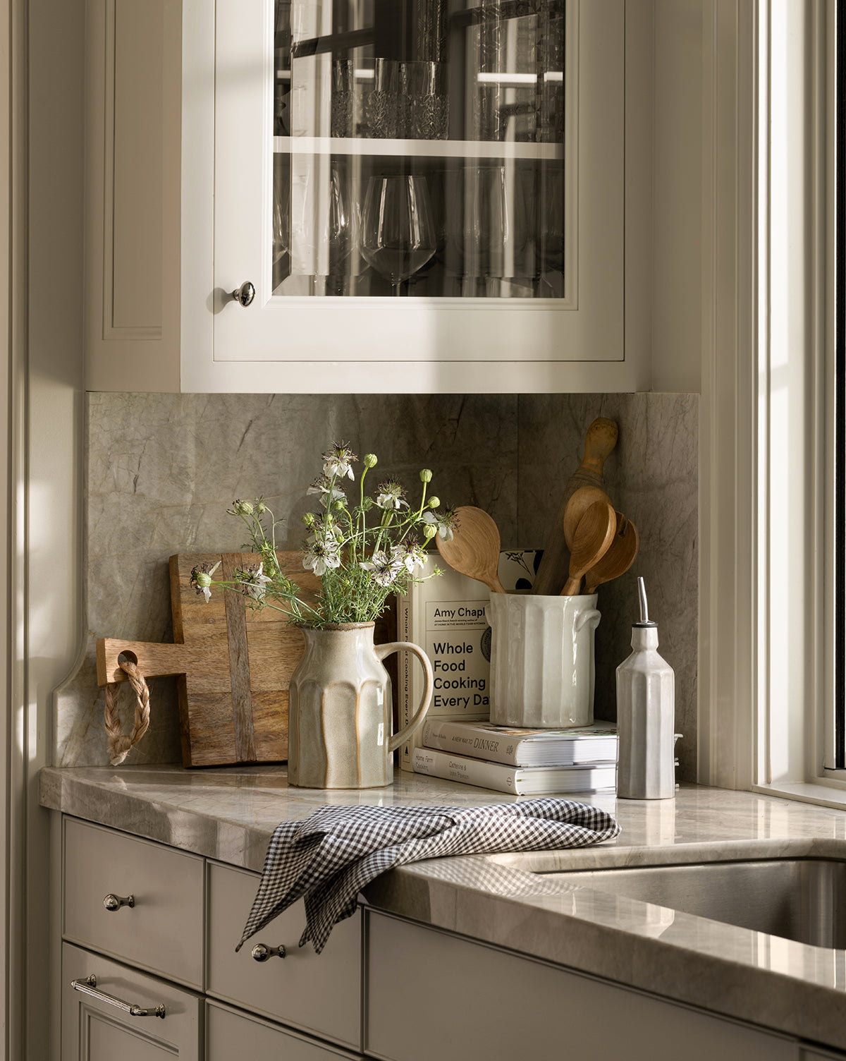 A sunlit kitchen countertop displays a pitcher of flowers, wooden cutting boards, cookbooks, utensils in a white container, stacked bowls, a towel, and the gray Emilia Cruet beside the glass soap dispenser by the sink.