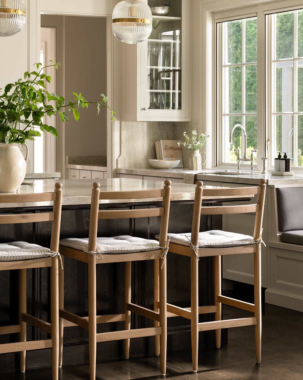 A bright kitchen features Romany Counter Stools at a marble island, a large potted plant, sunlit windows, built-in cabinetry, a farmhouse sink, and a window seat with a linen cushion by McGee & Co.