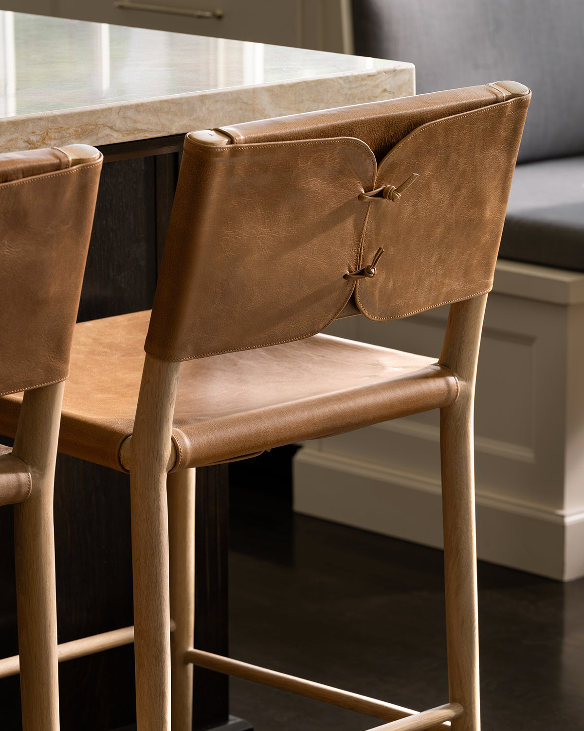 The Danson Counter Stool features a light wooden frame, tan leather seat and backrest with leather lacing, and minimalist design, shown beside a marble countertop in a stylish kitchen by McGee & Co.
