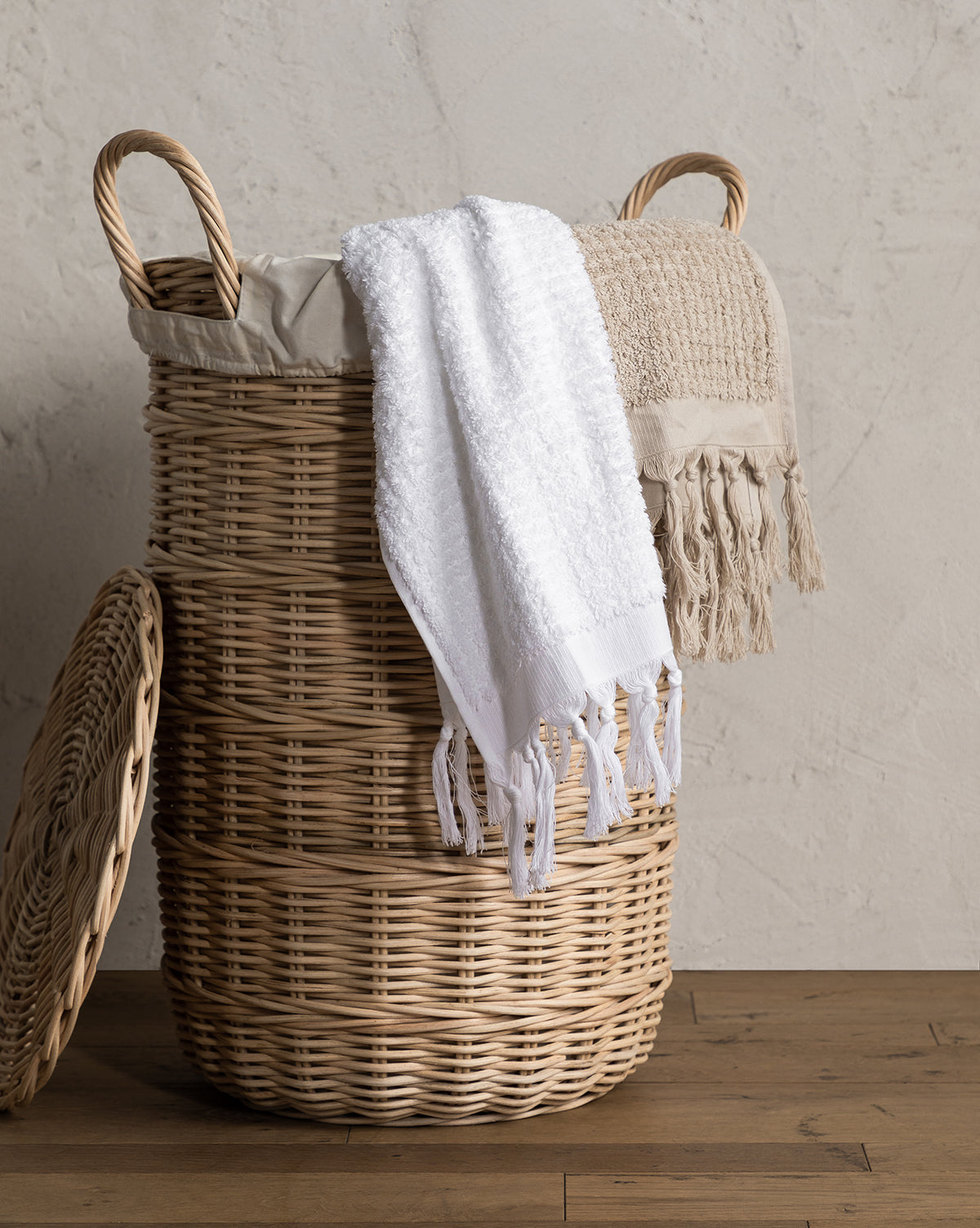 The Rattan Lidded Laundry Bin with two handles stands on a wooden floor, its lid propped against it and white and beige towels draped over the rim—an ideal stylish touch for any laundry room.