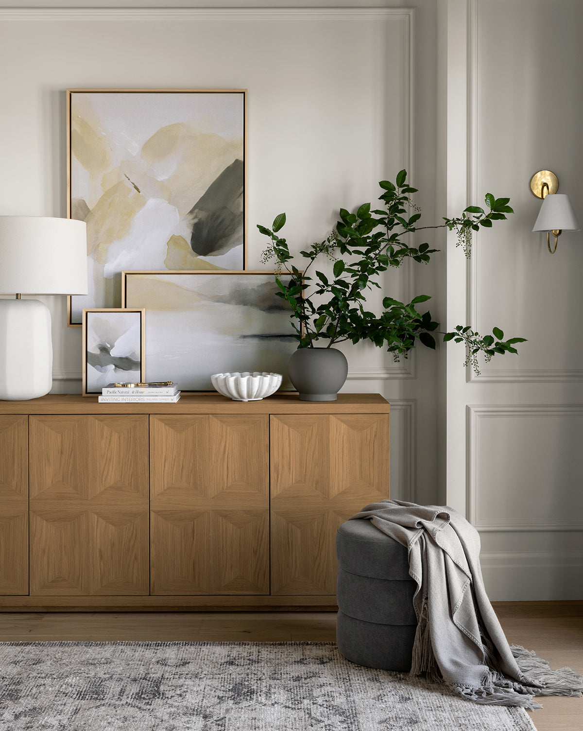 The Beacon Sideboard with a neutral finish anchors the room, topped with abstract art, a white lamp, books, a sculpted bowl, and a vase of leafy branches; nearby, a gray pouf and throw rest on a patterned rug in this calm space, by McGee & Co.