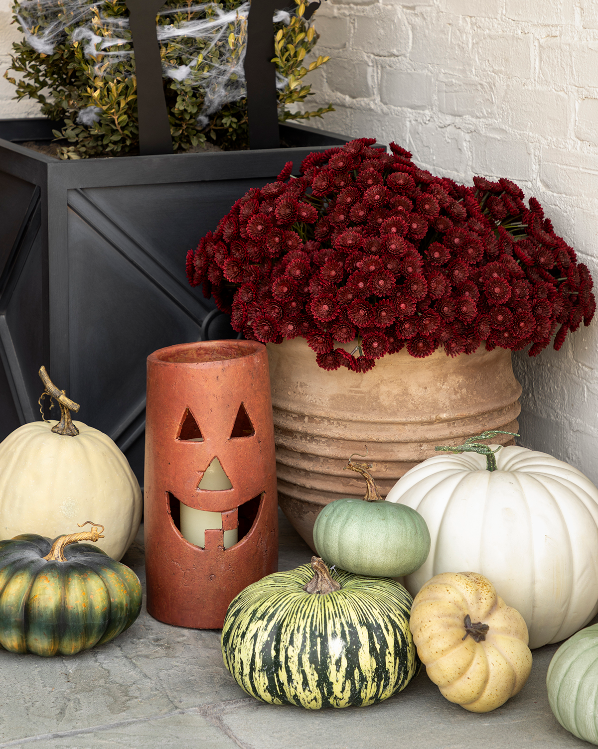 A fall porch display featuring white and green pumpkins, a terracotta jack-o-lantern candle holder, a black planter with greenery, and a pot of Faux Mum Drop-In blooms set against a light brick wall for inviting seasonal decor, by McGee & Co.