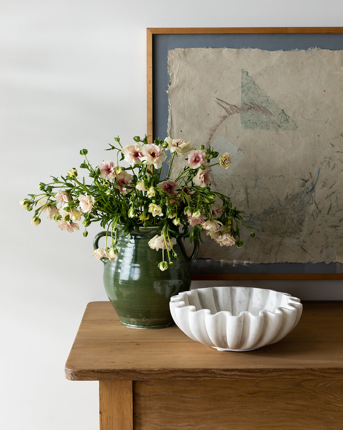 A green ceramic vase with pink and cream flowers sits on a wooden table beside the Fluted Marble Bowl, while a framed abstract drawing hangs on the wall behind them.