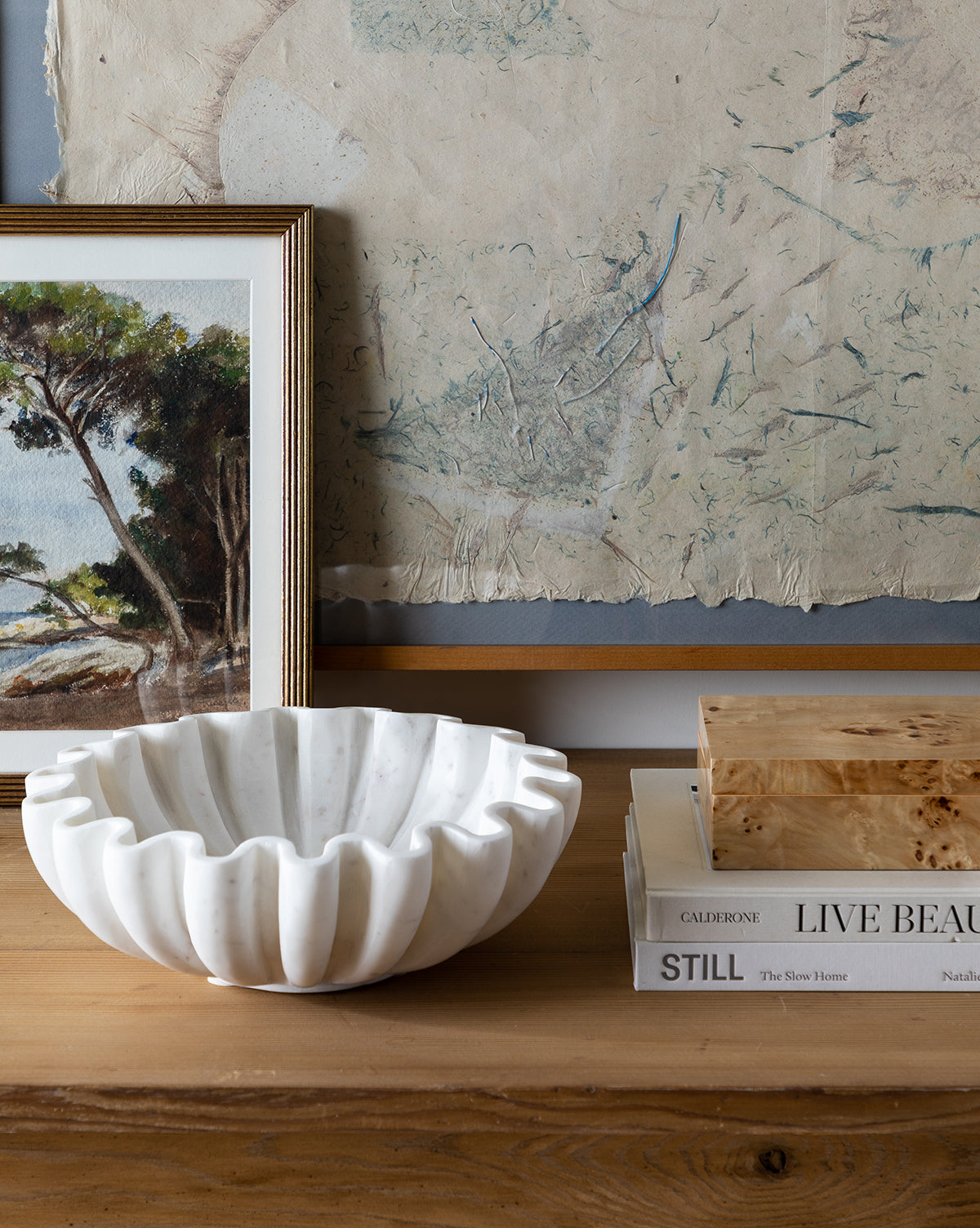 The Fluted Marble Bowl in white rests on a wooden surface next to stacked books and a wooden box, with a framed tree painting and abstract wall art in the background.