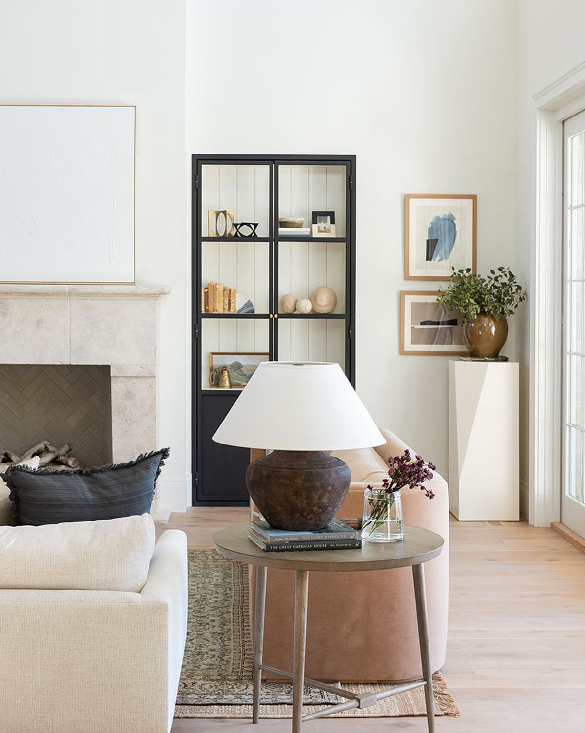 A bright living room with neutral tones features a stone fireplace, beige armchairs, a black pillow, the Tavin Side Table, a tall black bookshelf, framed wall art, and a plant on a white pedestal by a large window.
