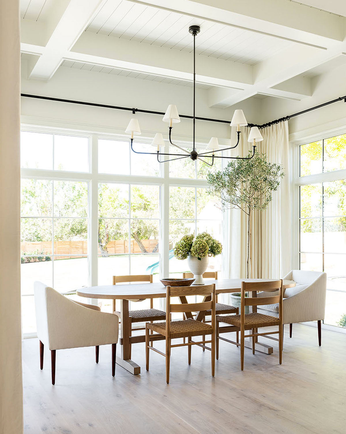 Bright dining room with large windows, light wood floor, the Giselle Oval Dining Table, mixed wooden chairs, two upholstered armchairs, potted plants, and a black modern chandelier hanging from a white beamed ceiling by McGee & Co.