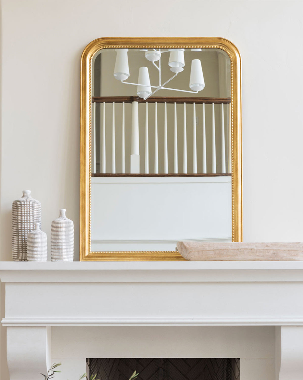 A Vienna Mirror with a classic molded border hangs above a white mantel decorated with three textured white vases and a rectangular tray. The gold-framed Vienna Mirror reflects a modern chandelier and vertical wooden railings, by McGee & Co.