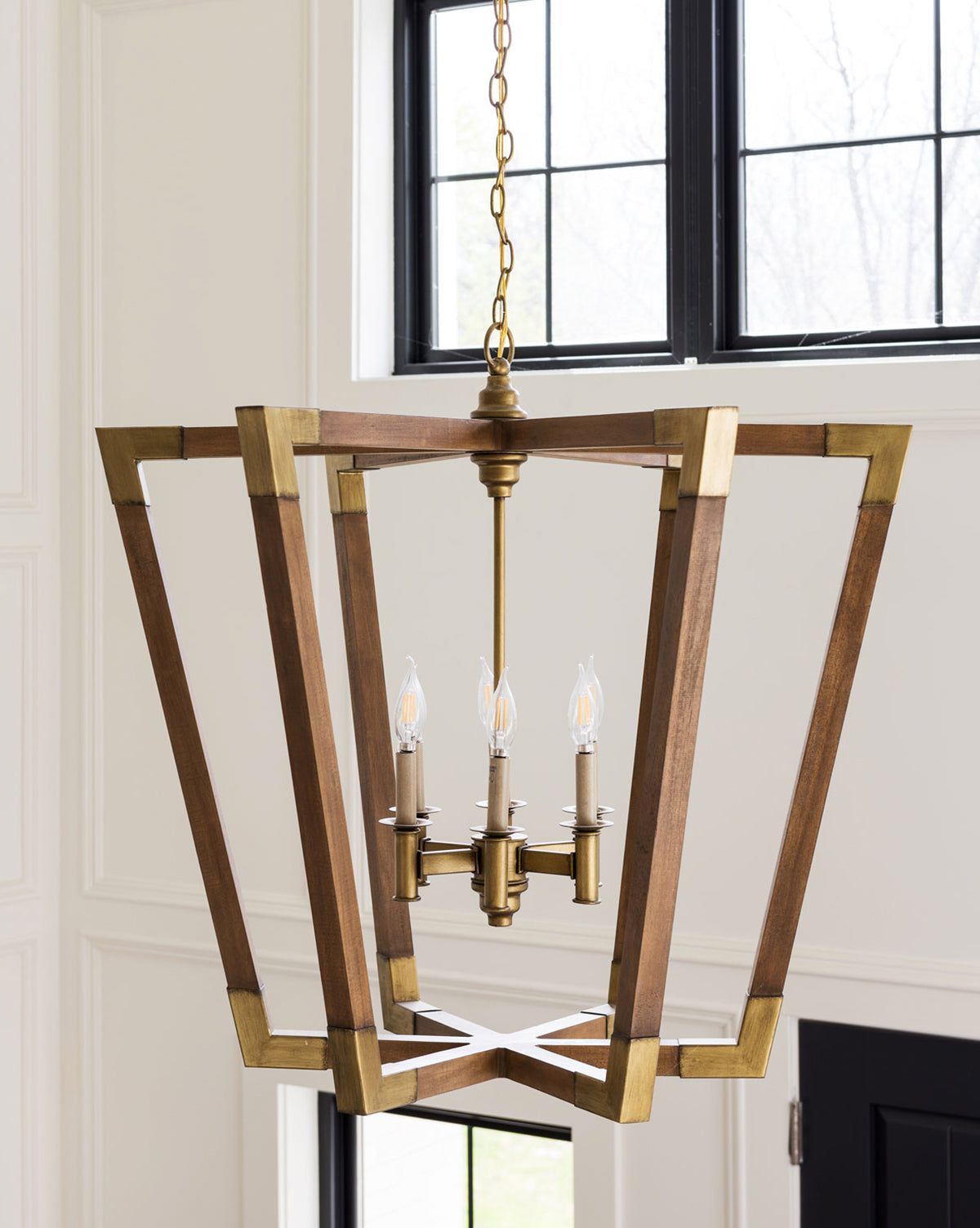 The Bastian Chandelier by Currey & Co. features a geometric chestnut and brass frame with four candle-style bulbs, adding modern elegance suspended near large black-framed windows. McGee & Co.
