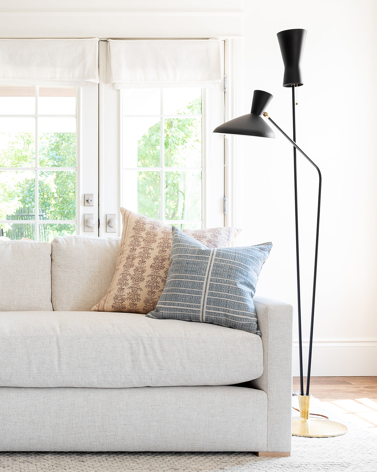 A modern living room with a light beige sofa, two patterned throw pillows, and the Austen Dual Function Floor Lamp. Large windows behind the setup allow natural light to stream in, showcasing green trees outside by McGee & Co.