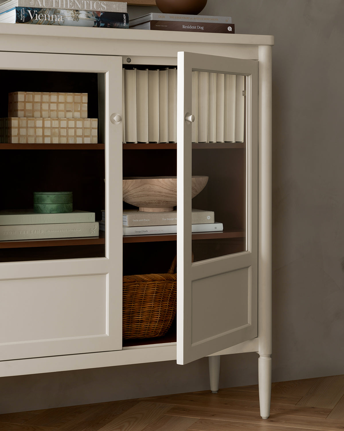 A bone white Highgate Cabinet with glass doors stands partly open, displaying shelves with books, a wooden bowl, a woven basket, and decorative boxes. Several books are stacked on top of this elegant Highgate Cabinet by McGee & Co.