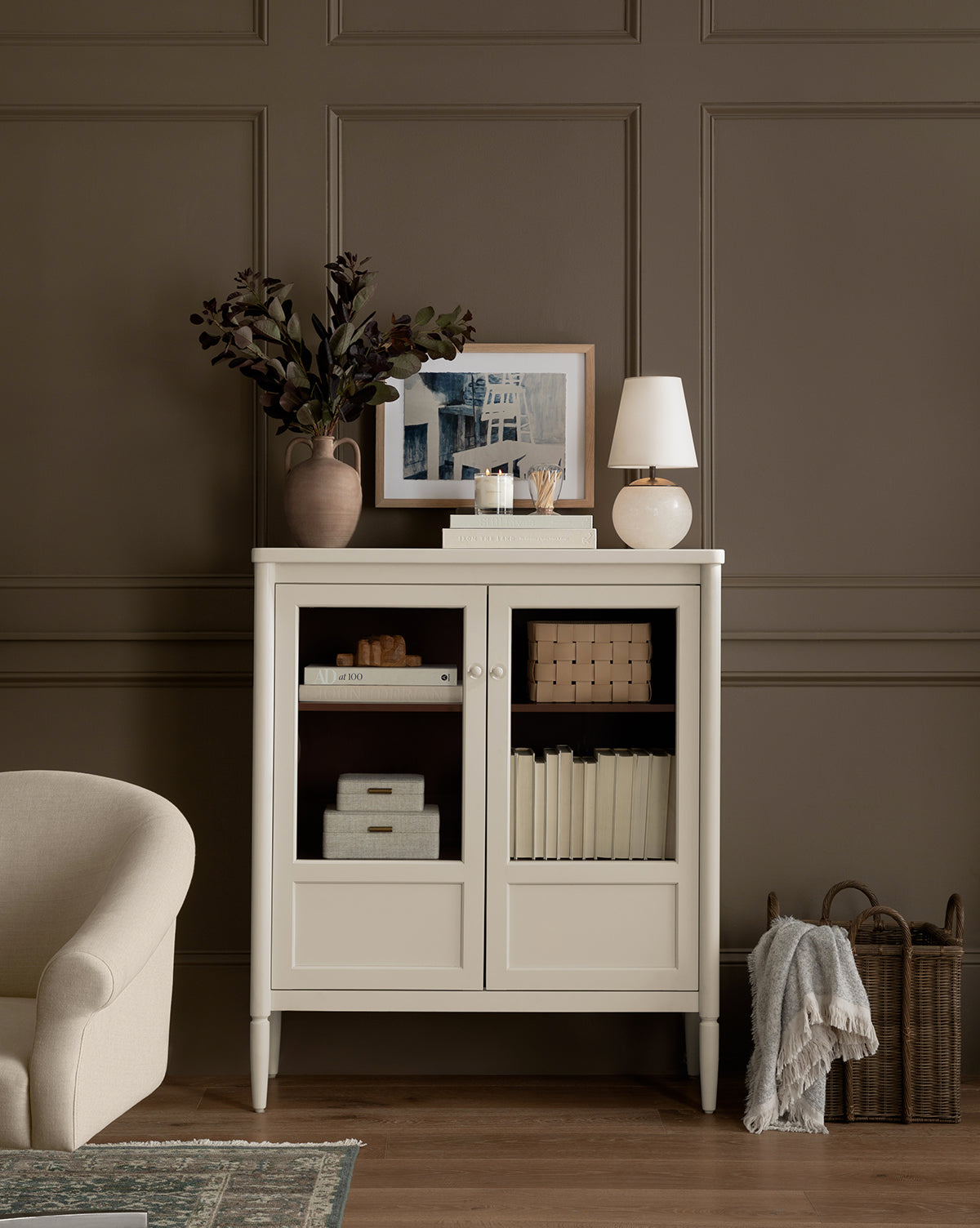 A beige cabinet with glass doors holds books and boxes. On top are a vase with branches, books, a wood frame displaying the Space Study art print, and a small lamp. A cozy armchair and blanket basket sit nearby against a brown paneled wall.