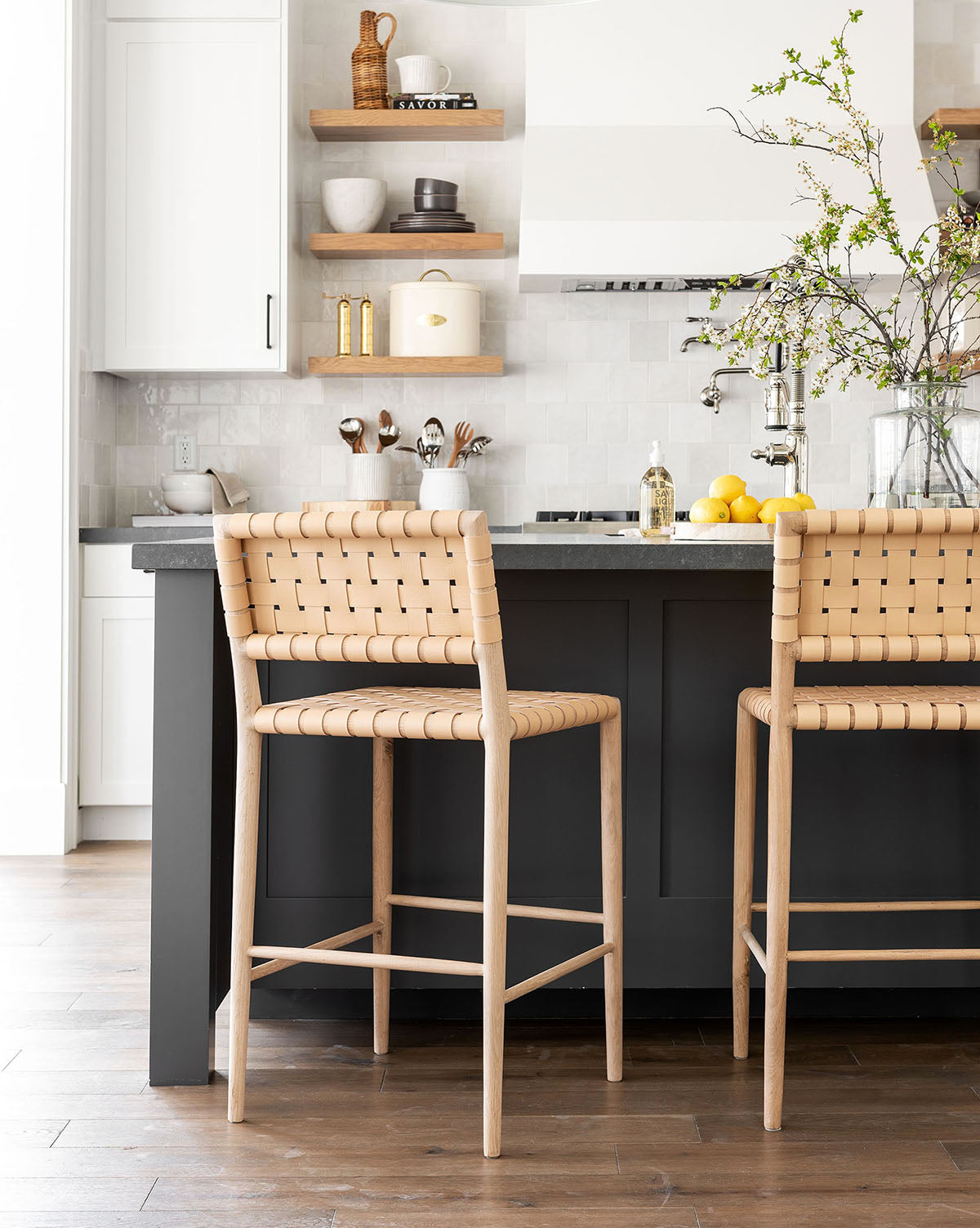 A modern kitchen features Greely Stools at a dark island, open wood shelves with dishes, a white backsplash, and a vase with greenery and lemons on the counter by McGee & Co.
