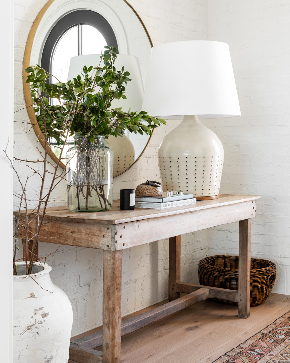 A rustic wooden table sits beneath the Solange Mirror—a minimalist piece with a thin stainless steel frame—adorned with a large ceramic lamp, a glass vase of greenery, stacked books, and a wicker basket below; a white vase rests on the floor, by McGee & Co.
