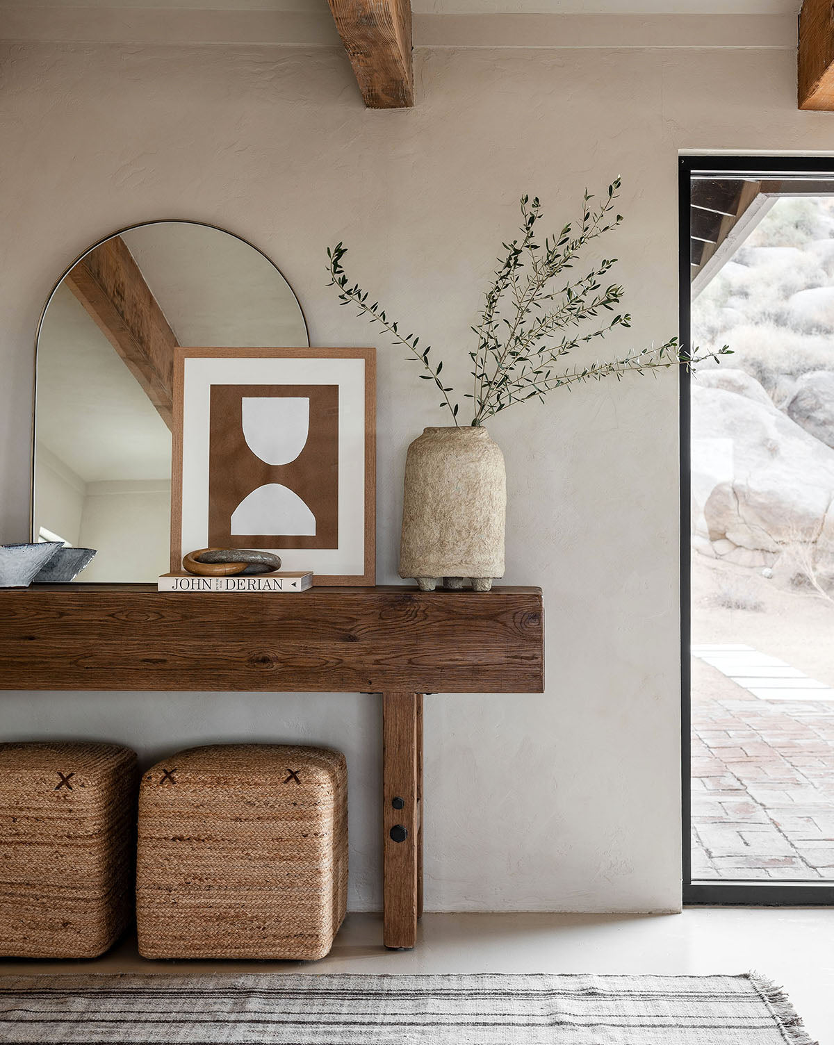A rustic wooden console table with the Wylie Arched Mirror, minimalist art, a textured vase with branches, and two woven ottomans below, set against a neutral wall near a door overlooking rocks outside by McGee & Co.