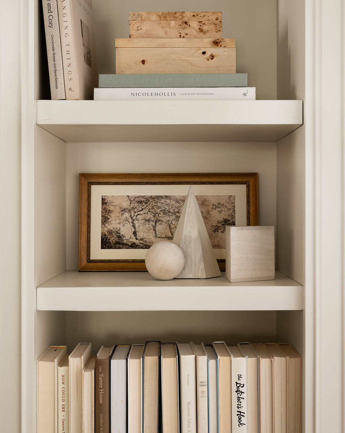 A white shelving unit displays stacked neutral books, a framed landscape photo, minimalist decor (wooden pyramid, sphere, rectangular block), and the Burl Wood Box with additional books neatly arranged on the bottom shelf.