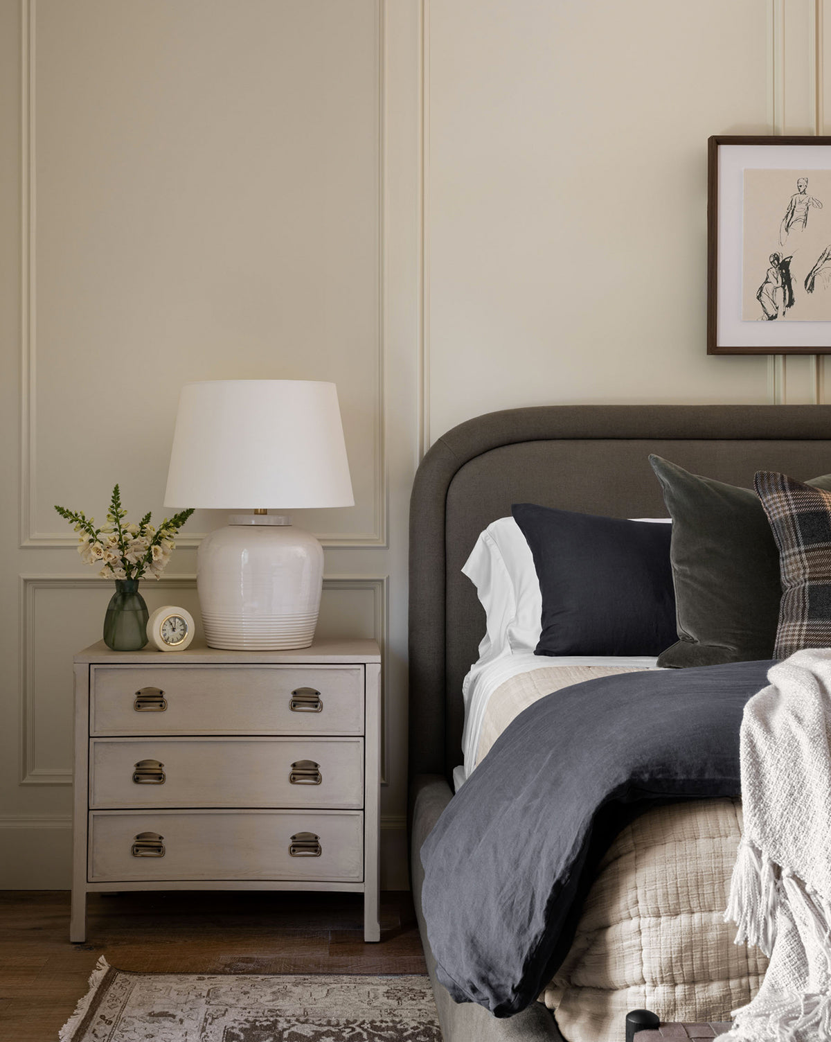A cozy bedroom with a modern gray upholstered bed, neutral bedding, and a nightstand featuring the Nora Table Lamp—ceramic base and linen shade—alongside a small clock and flowers against a light paneled wall by McGee & Co.