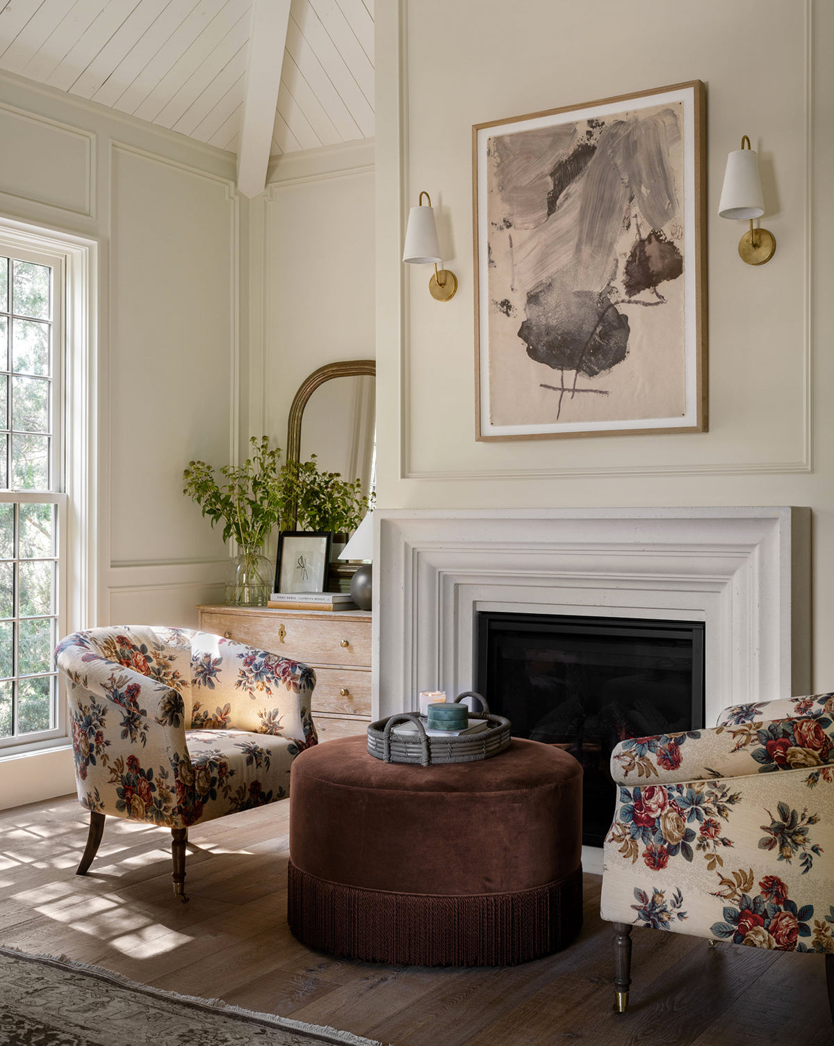 A cozy living room featuring two Swath 2 Sepia floral armchairs, a brown round ottoman, black fireplace, abstract art in a white oak frame above the mantel, wooden dresser with mirror, and a vase of greenery by a large window.