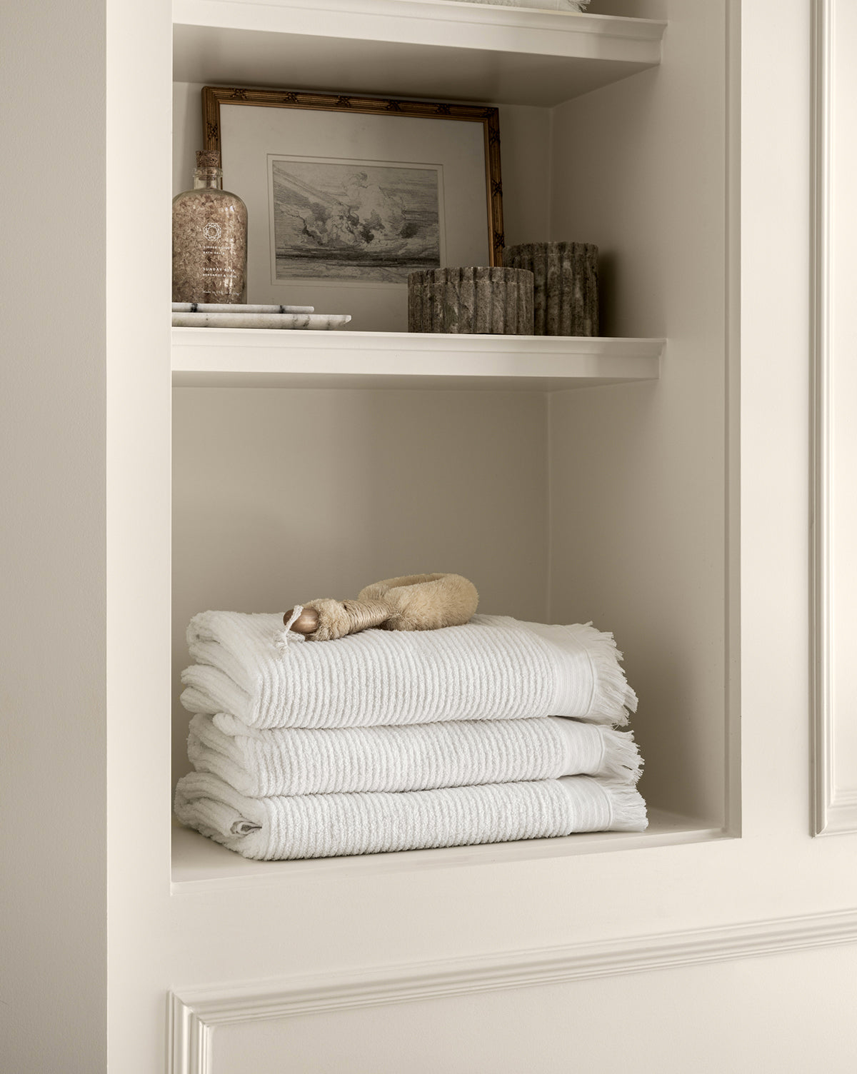 A shelf with three neatly folded white towels and a small beige plush toy is complemented by the Ethereal Clouds artwork in a gold wood frame, plus a decorative bottle and textured candle holders on off-white shelves.