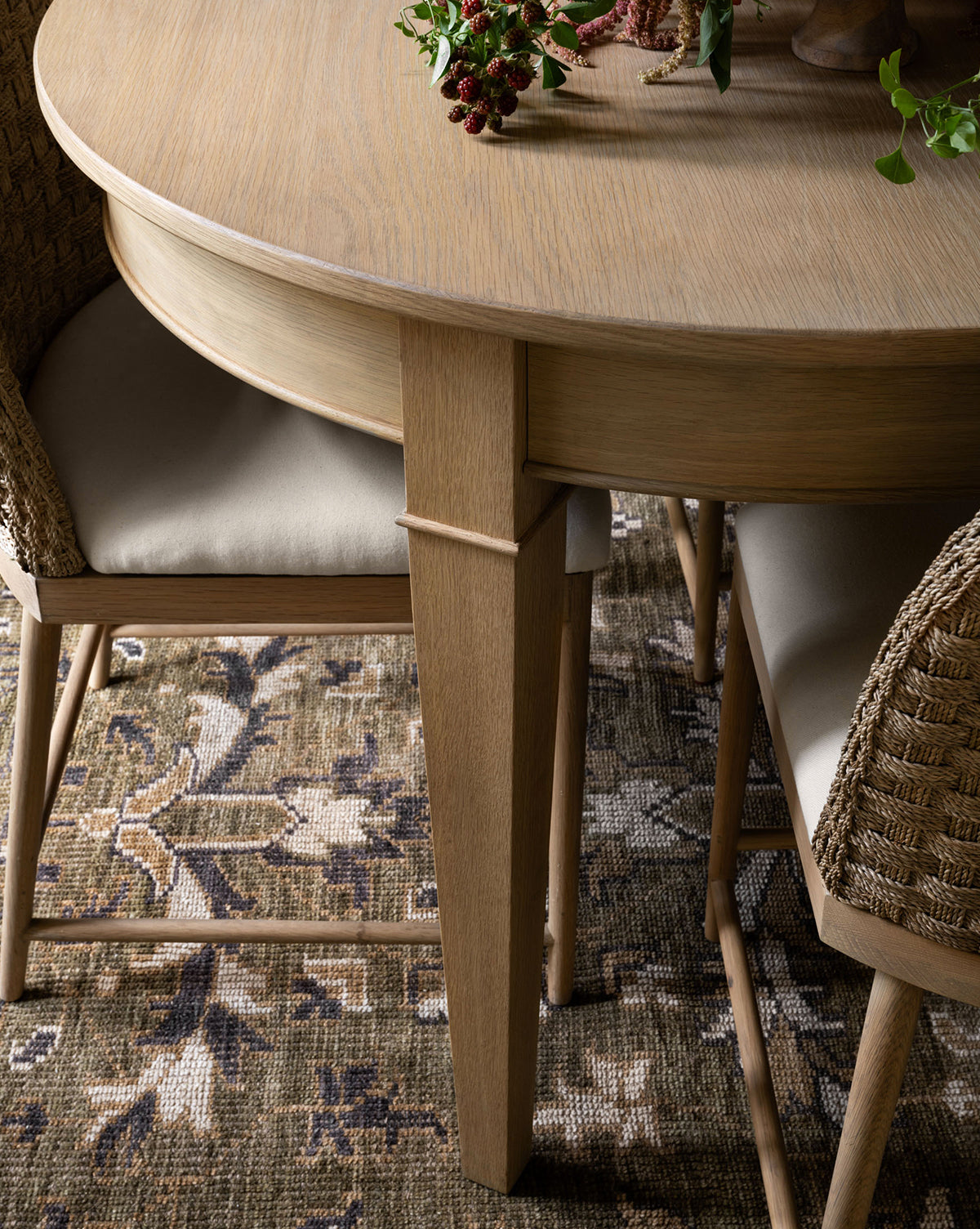 A round wooden dining table with woven chairs and beige cushions sits atop the Wayland Hand-Knotted Wool Rug. A small plant brings a lively accent to this cozy, inviting setting, by McGee & Co.