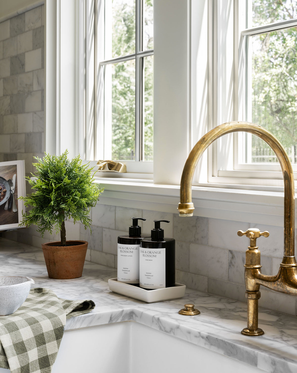 Close-up of a kitchen sink area with a brass faucet, soap bottles on a tray, and a checkered towel on marble. A 15.5" Cypress Faux Potted Topiary in a terracotta pot adds greenery beside large windows, by McGee & Co.