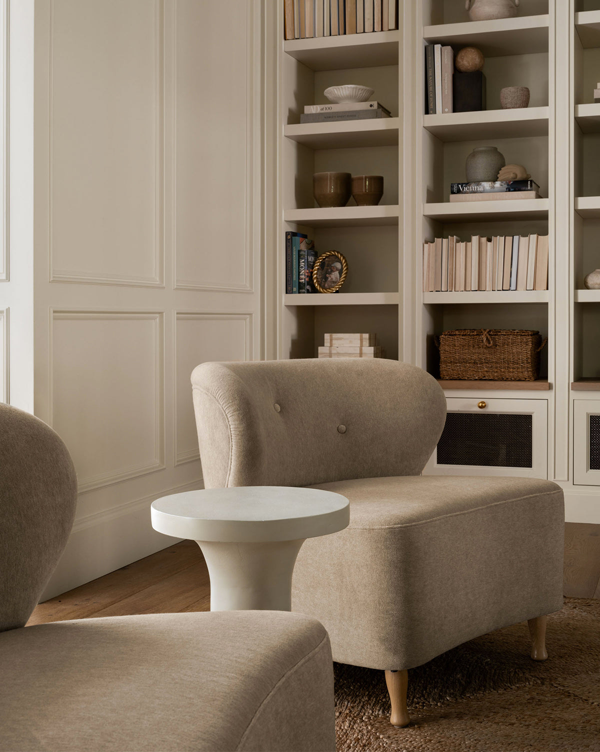 Two Carmella Slipper Chairs in beige sit with a small white round side table before built-in bookshelves filled with books and decor in a softly lit, minimalist room with wood flooring, by McGee & Co.