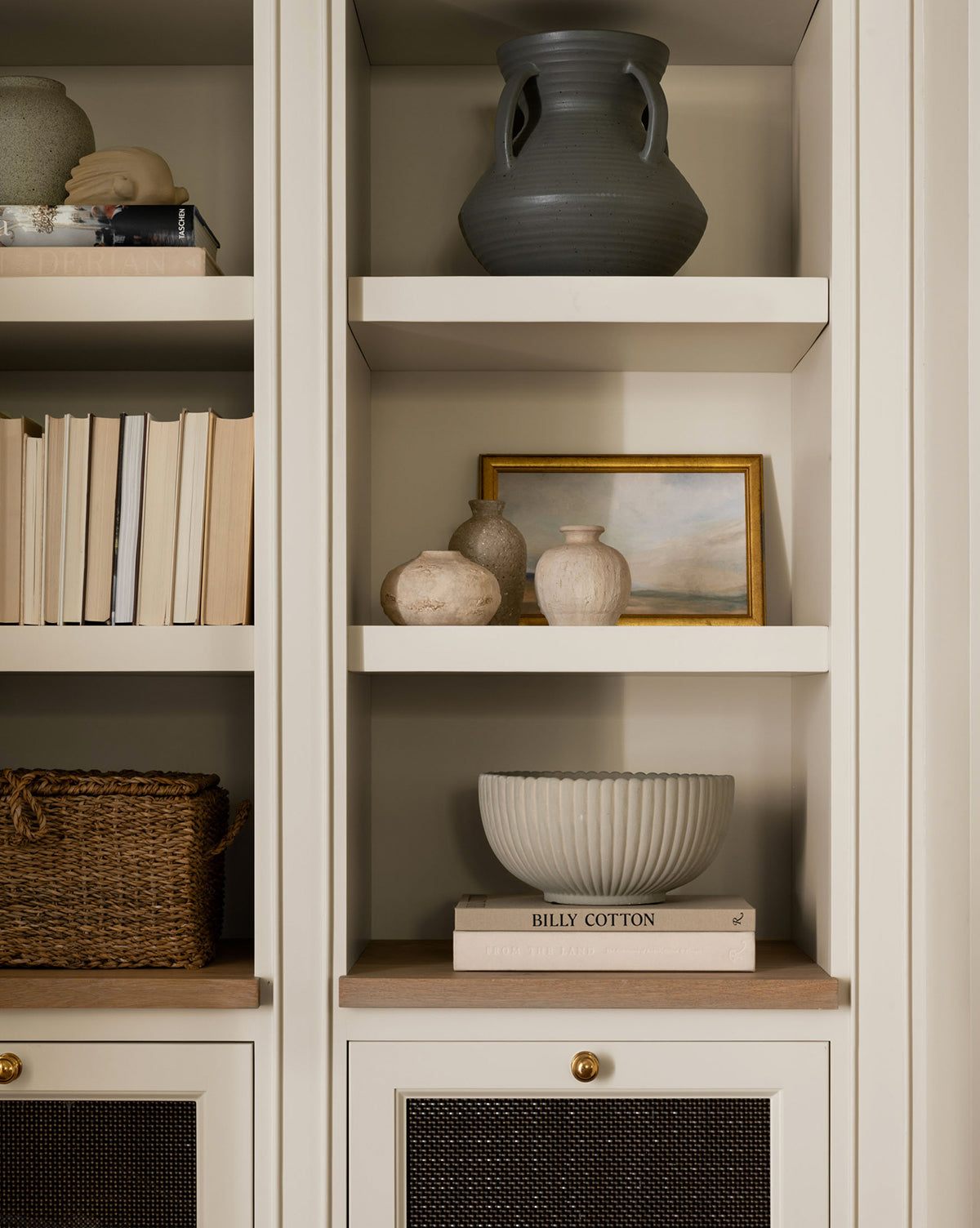 A neatly styled beige bookshelf displays books, a basket, Thalia Bud Vases (Set of 3), a large gray jug, a small framed painting, and a decorative bowl resting on two stacked books.