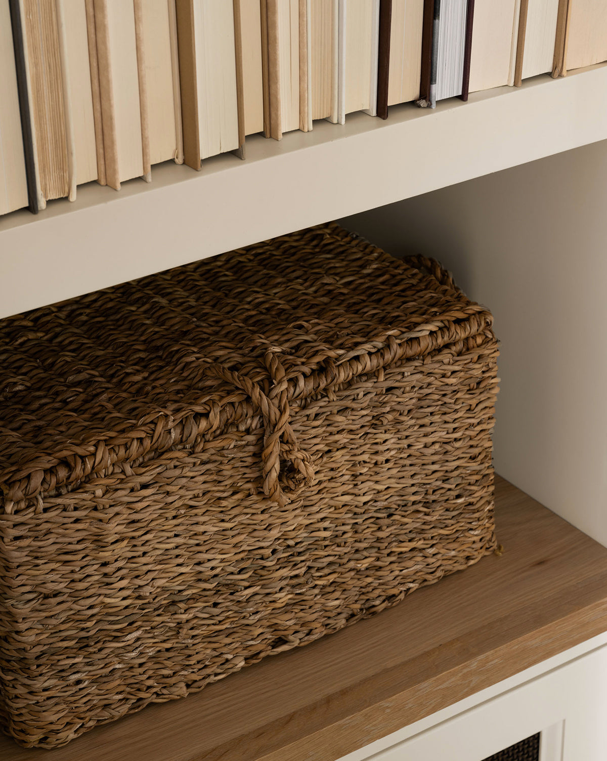 The Kya Seagrass Box, a rectangular hand-woven storage basket with a lid, rests on a wooden shelf below upright books with light-colored covers by McGee & Co.