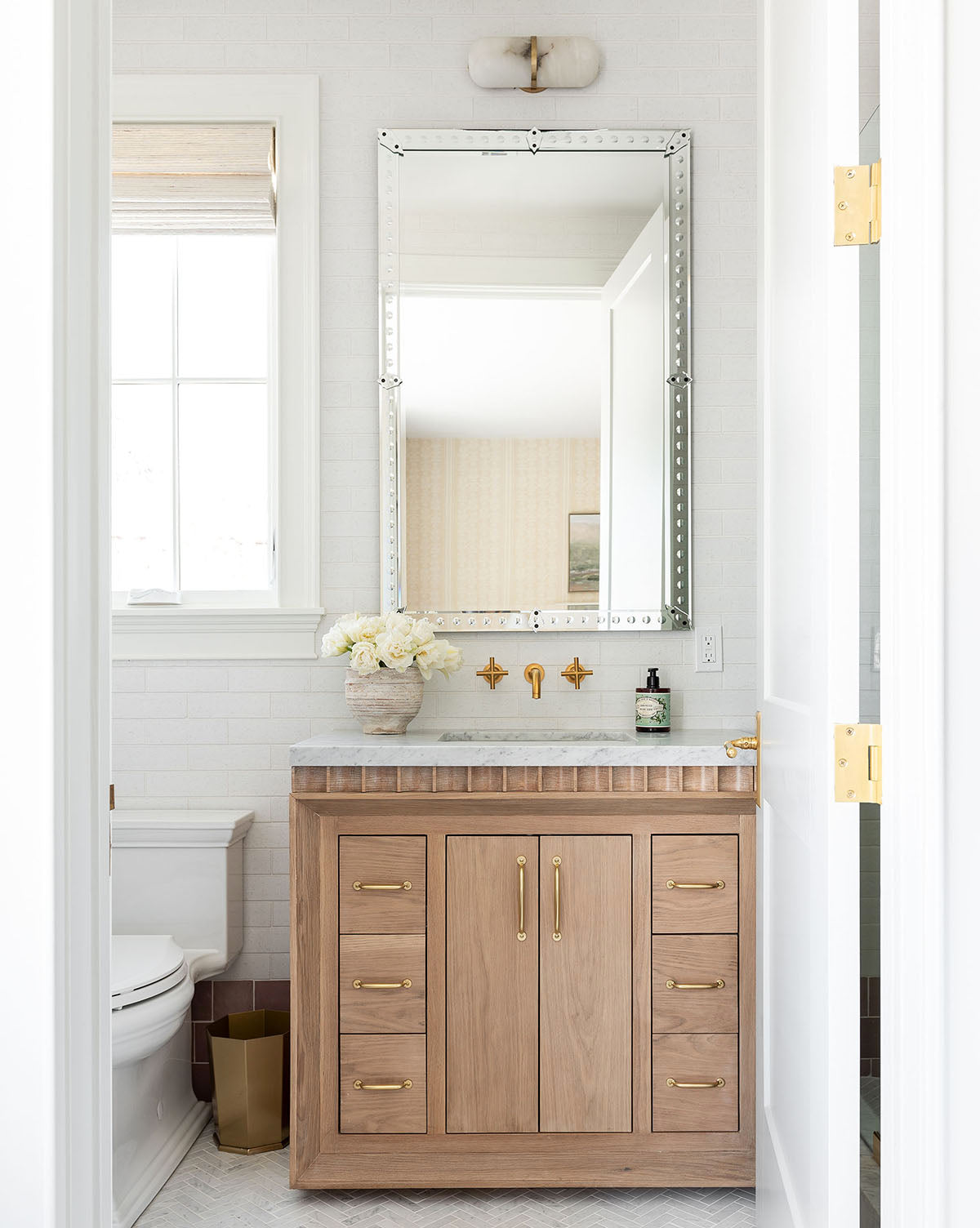 A bright bathroom showcases a light wood vanity with gold hardware, white countertops topped with the Truman Mirror, a wall-mounted gold faucet, a white flower vase, and a toilet beside the window, by McGee & Co.